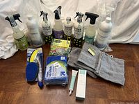 Front view of all cleaning supplies and accessories arranged on a wooden surface, showing spray bottles, sponges, washcloths, dish scrubber and skincare tube.