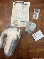 Top view showing the Oreck XL fabric shaver, power cord, user manual, and extra blades in a sealed plastic bag all laid on a wood surface.