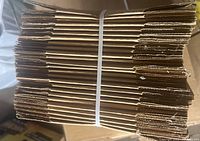 Side view of stacked flat-packed cardboard boxes secured with a white plastic strap.