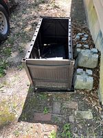 Photo of one black plastic storage box panel standing upright with wood grain texture, placed next to stacked concrete blocks on grass and brick ground.