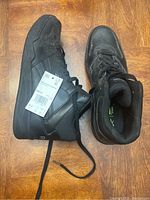 Pair of black Reebok high top shoes next to each other on wood floor, left shoe has tag attached with size and barcode.
