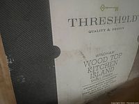 Close-up of box label showing the brand Threshold, product line Windham, and description: Wood Top Kitchen Island with Shell Finish, antique bronze hardware, casters, towel bar, and adjustable shelf.