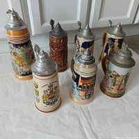 Seven steins arranged in two rows on white sheet, showing overall lot