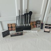 Full lot view showing makeup brush set in black holder, two Revolution cheek kit palettes, and boxed products including NARS blush, e.l.f. primer, Revolution brow pomade, No Dark Shadows enhancer, Lancome mascara, and Estee Lauder lipstick.