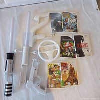 Photo showing the full lot of Nintendo Wii accessories and games arranged on white backdrop, including Star Wars lightsabres, Wii steering wheel, gun controller, sensor bar holder, and 6 game cases.
