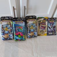 Photo showing all five sealed Pokemon card products arranged side-by-side, including three starter set tins and two booster packs.