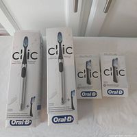 Photo showing two boxed Oral-B Clic manual toothbrushes and two boxes of replacement brush heads, all in original sealed packaging.