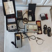 Overview of vintage diagnostic meters and tools laid out on white surface, showing Amprobe AM-3 multimeter, voltage supply, thermometer, amp probes, dial indicator, and other devices.