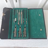 Open drafting set case showing seven metal drawing instruments placed on green felt interior.