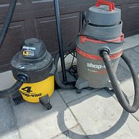Photo shows two wet/dry vacuum cleaners side by side: a yellow 4 gallon Shop-Vac with black top and hose, and a larger gray 8 gallon Shop-Vac with red trim and hose attached, both on a paved surface outdoors.