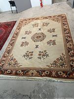 Full view of the cream wool area rug with floral and geometric patterns spread on concrete floor showing the entire rug including its fringe and pattern layout.