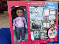 Front view of the boxed American Girl doll with two outfits and accessories visible in the packaging window.