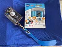 Photo front view of VTech KidiZoom PrintCam box and Franklin NHL hockey stick with two NHL balls on blue background.