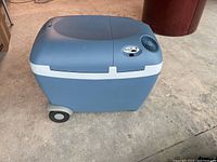 Side angle view of blue and gray Mobilcool cooling and warming box showing wheels and part of control panel on top.