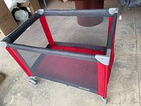 Photo of Avalon fold up pack'n'play from one angle showing red fabric, gray mesh sides, and gray padded top with wheels on one side.