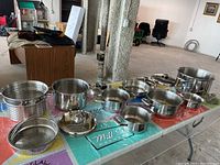 Top-down view of pot set including vegetable steamer insert, six pots, and four lids arranged on colored tablecloth.