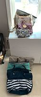 View of eight assorted ladies tote bags and handbags stacked on a white dresser and on the floor nearby. Visible styles include printed fabric, stripes, and solid colors.