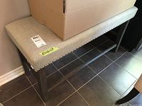 Photo of upholstered bench showing beige fabric padded seat with silver nailhead trim and wood veneer legs.
