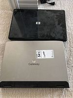 Two laptops laid closed on the carpet: one black HP laptop on top, one silver Gateway laptop below