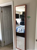 Full view of a full-length rectangular wall mirror with reddish-brown metal frame mounted on a wall next to a doorway.