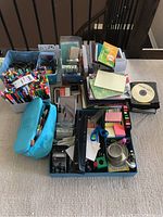 Photo of assortment of pens, markers, scissors, notebooks, sticky notes, CDs and organizers arranged on floor carpet.