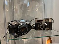 Olympus OM10 camera with lens next to Dignette 300 camera on glass shelf