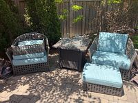 Two wicker patio chairs and one ottoman with cushions positioned around a table, covers draped on ground