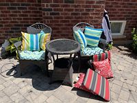 Overview of two dark brown wicker armchairs flanking round wicker side table with glass top and assorted cushions