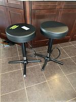 Two black adjustable height bar stools with round padded seats and black metal bases with footrests, placed on tiled floor in front of wooden cabinets.