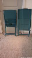 Rear view of two folded ocean green nylon folding chairs showing metal frames and handles on backrests.