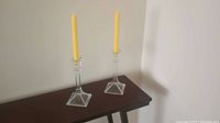 Photo of two clear glass candle holders each with a tall yellow candle standing on a dark wooden surface against a plain white wall.