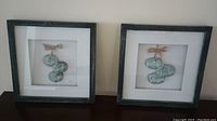 Two dark gray square frames each containing a set of blue-green hanging stones with white inspirational sayings, behind glass.