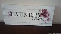 Photo of wooden laundry room plaque with floral design on a white paneled background.