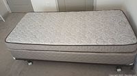 Twin mattress and boxspring set on wheeled base against wall on carpeted floor.