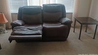 Brown leather reclining loveseat with one footrest extended, placed in a carpeted room with side table visible.