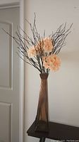 Tall brown rectangular glass floor vase holding peach artificial flowers and multiple long brown decorative twigs, against a beige wall and dark surface.
