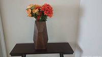 Tall wood vase with geometric faceted sides placed on a dark wood table against a white wall, holding artificial orange and yellow flowers.