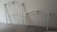 Photo showing white antique finish twin headboard and footboard, both with curved metal detailing and round decorative elements, in a carpeted room against a plain wall.