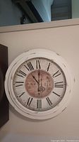 Large round wall clock with white distressed frame hanging on wall, featuring Roman numerals and classic black clock hands