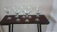 Photo showing eight assorted clear glass wine glasses with long stems placed on a dark wooden table against a plain light colored wall.