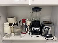 Shelf of small kitchen appliances including a Black and Decker blender, Sunbeam toaster, GE hand mixer, metal fondue pot, cheese grater, and small chopper appliance