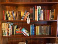 Two wooden shelves filled with books, mostly hardcover, arranged vertically and some horizontally. Books vary in size, color, and age, showing vintage and antique characteristics.