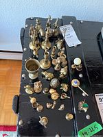 Top-down view of various miniature brass figurines including cups, animals, and small decorative items arranged on a black surface.