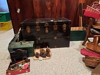 Front view of a large black metal chest trunk with brass locks and latches, showing riveted metal edges and hardware, sitting on carpeted floor with some small vintage items surrounding it.