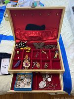 Full view of open beige jewelry box showing red velvet compartments with assorted pendants, pins, and charms inside.