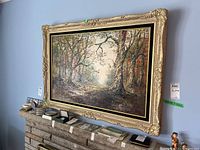 Framed oil painting of a forest path with visible signature and detailed tree bark texture.