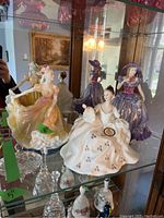 Three Royal Doulton figurines displayed in a glass cabinet, showing the distinct dresses and postures of each figure.