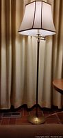 Full view of the brass floor lamp with cream-colored fabric shade, showing the lamp in front of a curtain.