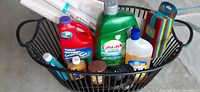 Photo of cleaning supplies in black wire basket showing detergent bottles, small oil bottle, cleaning sponges, brush and rolled white sheets.