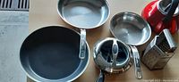 Top-down view showing a non-stick frying pan, a smaller stainless steel pan, a double boiler insert, a small stainless steel bowl, and a box cheese grater on a wood surface.